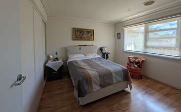 This cozy bedroom features a neatly made double bed, wooden floors, a nightstand, a large window with blinds, artwork above the bed, and a plush armchair.