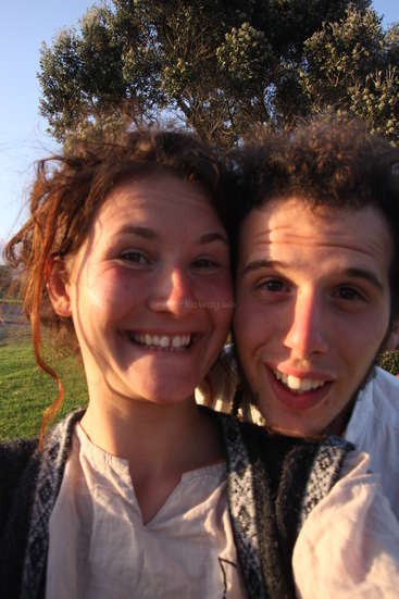 Das Bild zeigt ein Selfie von einem Mann und einer Frau mit lockigem Haar, die in die Kamera lächeln, vor einem Hintergrund aus Bäumen und Gras an einem sonnigen Tag.