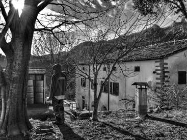 Das Bild zeigt eine ruhige Schwarz-Weiß-Szene mit einer Person, die in einem Garten steht, umgeben von Bäumen und einem Haus, mit einer Vogelfutterstelle und anderen Gegenständen in der Nähe.