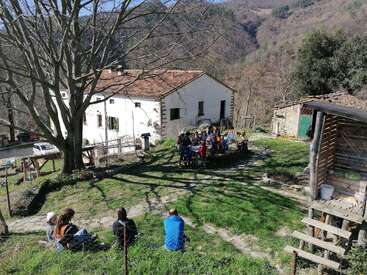 A group of people gathers outdoors near a rustic house, surrounded by hills and trees. Some sit on the grass, others at a large table, enjoying nature.