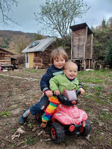 Two young children joyfully ride a red toy quad bike outdoors, surrounded by rustic wooden cabins, trees, and hills, with cloudy skies overhead and muddy ground below.
