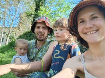 A happy family of four sits outside on a sunny day, smiling and enjoying the nature around them, surrounded by greenery and tall trees.