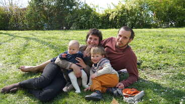 A happy family of four relaxes on a green grassy lawn. The parents and two young children smile and pose outdoors on a sunny day.