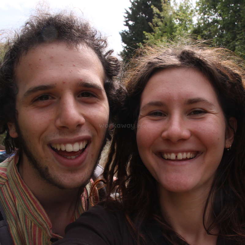 Das Bild zeigt ein Selfie von einem Mann und einer Frau mit dunklen Haaren, die in die Kamera lächeln, vor einem Hintergrund aus Bäumen und einem hellblauen Himmel.