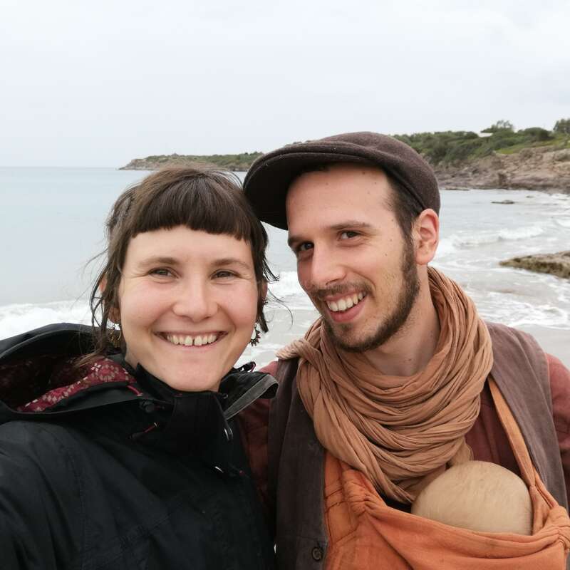 A smiling couple stands on a beach, the man carrying a baby in a sling. The background shows ocean waves, rocky shore, and cloudy sky.