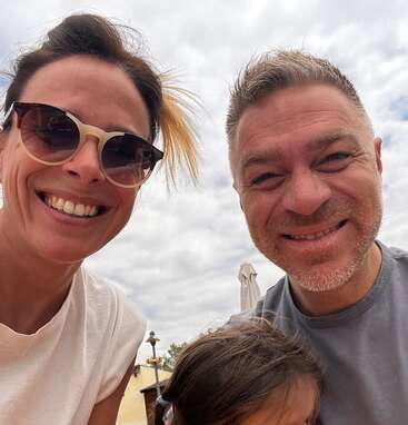 A smiling woman in sunglasses, a grinning man, and a child partially visible at the bottom pose together outdoors under a cloudy sky, enjoying their time.