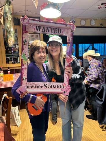 Zwei Frauen posieren gemeinsam auf einer Party und halten einen dekorativen „WANTED“-Rahmen in den Händen. Eine von ihnen hält eine Spielzeuggitarre in der Hand, Cowboyhüte und Western-Dekorationen prägen den lebhaften Hintergrund.