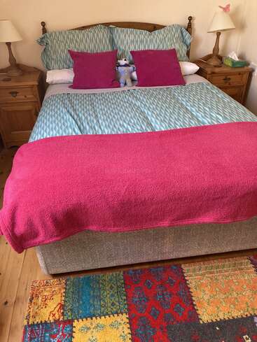 Auf diesem Bild ist ein ordentlich gemachtes Bett mit rosa-blauer Bettwäsche, zwei rosa Kissen, einem Stofftier, Nachttischen aus Holz, Lampen und einem bunt gemusterten Teppich zu sehen.