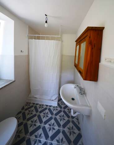 Este cuarto de baño cuenta con una pequeña ducha con cortina blanca, suelo de baldosas estampadas, lavabo montado en la pared, armario de madera, ventana y una única bombilla a la vista.