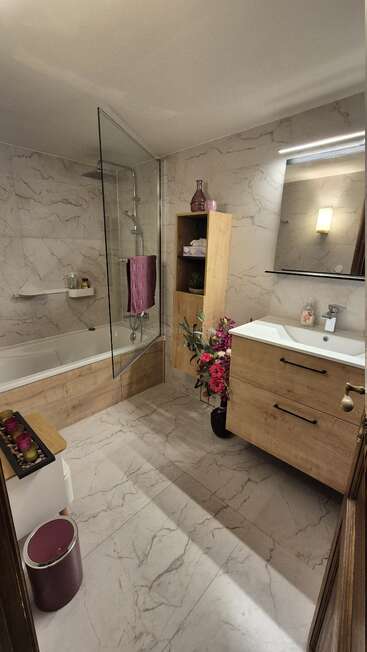 L'image montre une salle de bains moderne avec une baignoire blanche, une douche en verre et un meuble-lavabo en bois, dont les murs et le sol sont recouverts d'un carrelage ressemblant à du marbre.