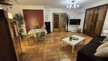 Ce salon chaleureux est agrémenté d'un sol en terre cuite, d'un canapé noir, d'une table basse, d'une table à manger ronde, d'une télévision, de grandes fenêtres, de plantes d'intérieur et d'un éclairage chaleureux.