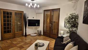Ce salon chaleureux est agrémenté d'un canapé noir, d'une table basse blanche, d'une télévision, de portes vitrées, de plantes d'intérieur, de touches de bois et d'un éclairage tamisé, créant ainsi une ambiance moderne et accueillante.