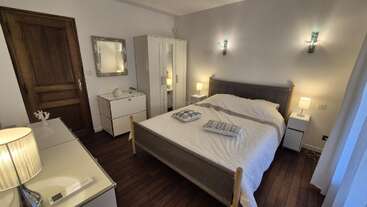Um quarto aconchegante e moderno com piso de madeira, uma cama estofada em cinza, móveis brancos, luminárias, arandelas, um espelho grande e uma luz natural suave que entra pelas cortinas.