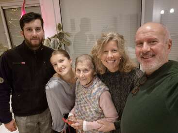 Cinco pessoas de diferentes idades estão bem juntas dentro de casa, sorrindo felizes para uma foto em grupo. No fundo, dá pra ver janelas, plantas e uma iluminação suave de ambiente.