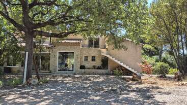 Uma charmosa casa de pedra cercada por árvores frondosas. Possui janelas amplas, uma escada externa sombreada por galhos maduros e um ambiente sereno e natural, com a luz do sol entrando suavemente.