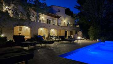 Uma elegante villa de pedra fica lindamente iluminada à noite, com pátios em arco, poltronas confortáveis e uma piscina azul cintilante, cercada por uma vegetação exuberante.