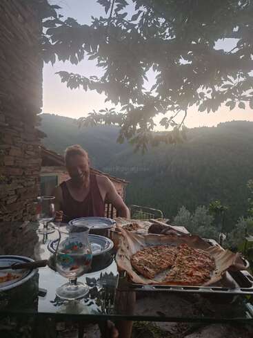 Ein Mann genießt eine Mahlzeit im Freien an einem rustikalen Tisch, umgeben von Bergen und Bäumen bei Sonnenuntergang. Selbstgemachte Pizza, Getränke und Brot schaffen eine gemütliche Atmosphäre.