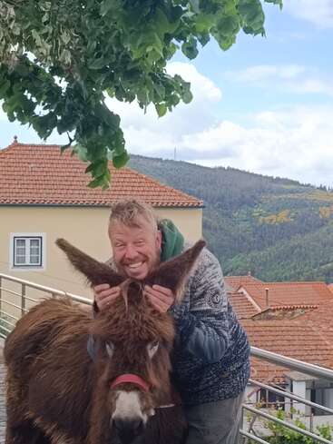 Ein lächelnder Mann hält spielerisch die Ohren eines Esels und posiert fröhlich im Freien. Dahinter liegen rote Ziegeldächer, grüne Hügel und ein belaubter Baum über ihnen.