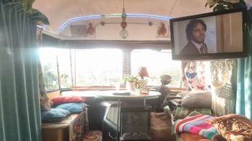 O interior aconchegante do ônibus tem almofadas coloridas, uma mesa redonda com plantas, cobertores estampados e uma TV mostrando um cara de terno, criando um clima caseiro.