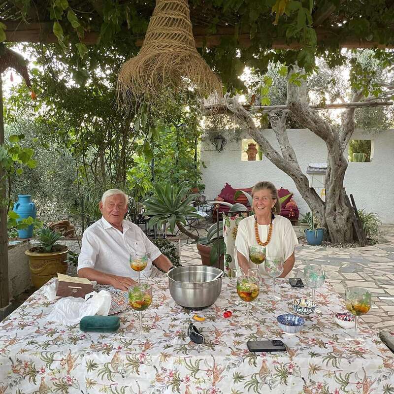 The image depicts a serene outdoor setting, featuring a couple seated at a table adorned with a vibrant tablecloth, surrounded by lush greenery and a rustic pergola.
