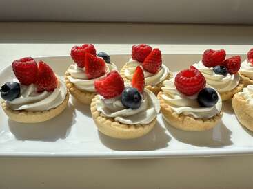 Six délicieuses tartelettes au fourrage crémeux, chacune garnie de framboises, de myrtilles et de fraises fraîches, sont disposées soigneusement sur un plateau rectangulaire blanc, à la lumière du soleil.