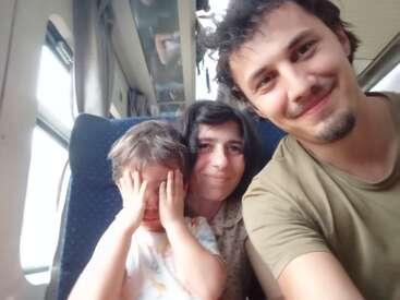 Um homem, uma mulher e uma criança estão sentados juntos em um trem. O homem sorri, a mulher parece satisfeita e a criança cobre o rosto.