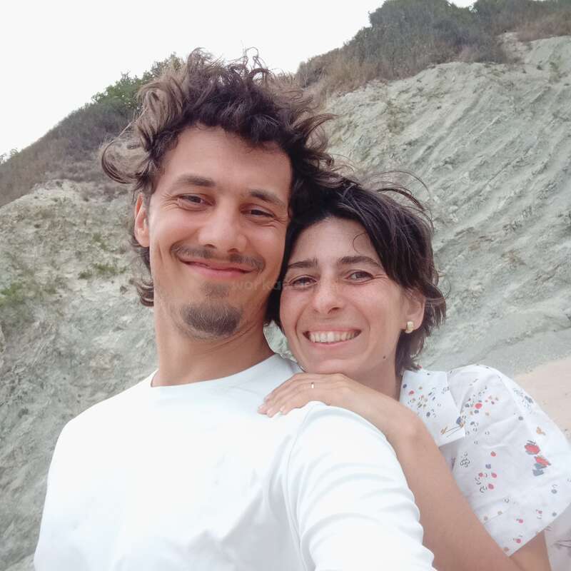 Um casal sorridente tira uma selfie ao ar livre, bem pertinho um do outro, em frente a uma encosta rochosa. Os dois parecem felizes, curtindo um momento descontraído e alegre juntos.