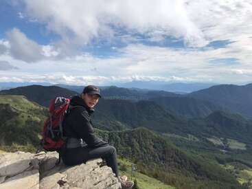Ein Mann sitzt mit einem roten Rucksack und schwarzer Kleidung auf einem Felsvorsprung und blickt auf eine Berglandschaft vor einem wolkenverhangenen blauen Himmel.