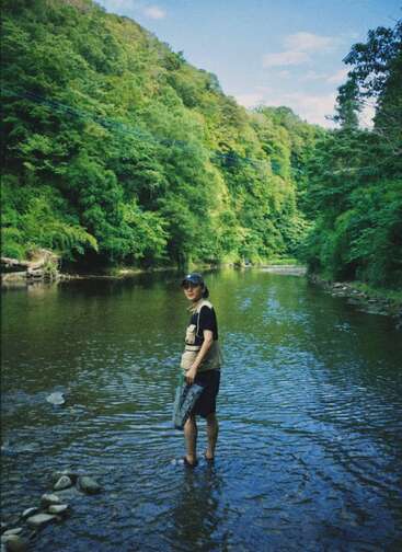 Eine Person steht in einem Fluss, trägt eine Schwimmweste und hält ein Netz in der Hand. Im Hintergrund ist ein bewaldeter Hügel an einem sonnigen Tag zu sehen.