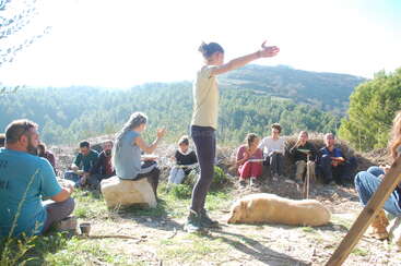 Das Bild zeigt eine Gruppe von Menschen, die in einem Feld auf dem Boden sitzen, eine Person steht und gestikuliert, umgeben von einem Hund und einer ruhigen Landschaft.