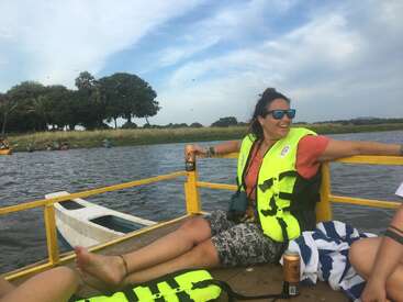 Une femme portant un gilet de sauvetage fluo se détend sur un bateau, souriante, avec des lunettes de soleil, en tenant un verre. Il y a de l'eau, des arbres et des gens qui font du kayak en arrière-plan.
