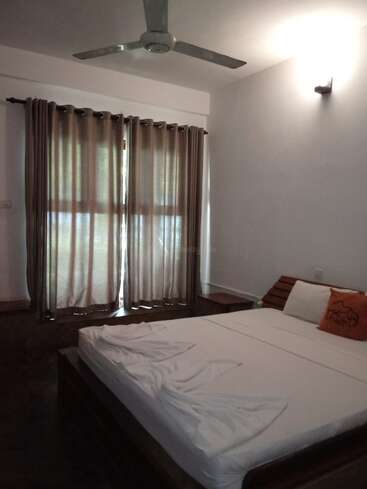 Une chambre simple comprend un lit double avec des draps blancs et un oreiller orange, de grandes fenêtres avec des rideaux bruns, un ventilateur de plafond, des murs et des éclairages minimalistes.