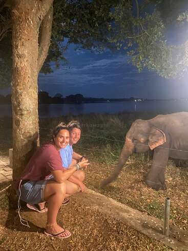 Two women crouch and smile under a tree at night beside a baby elephant reaching through a fence, with a serene lakeside landscape illuminated behind them.