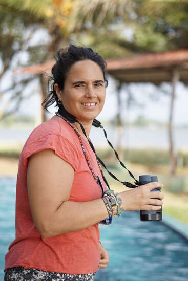 Une femme se tient à l'extérieur, au bord de l'eau, et sourit à l'appareil photo. Elle porte une chemise corail, des bracelets colorés et tient des jumelles, ce qui suggère qu'elle est en train d'observer les oiseaux ou de faire du tourisme.