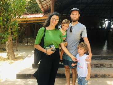 Une famille de quatre personnes se tient à l'extérieur. Le père et la mère tiennent chacun un enfant dans leurs bras. Ils sont habillés de façon décontractée, souriants, avec des arbres et un bâtiment rustique derrière eux.