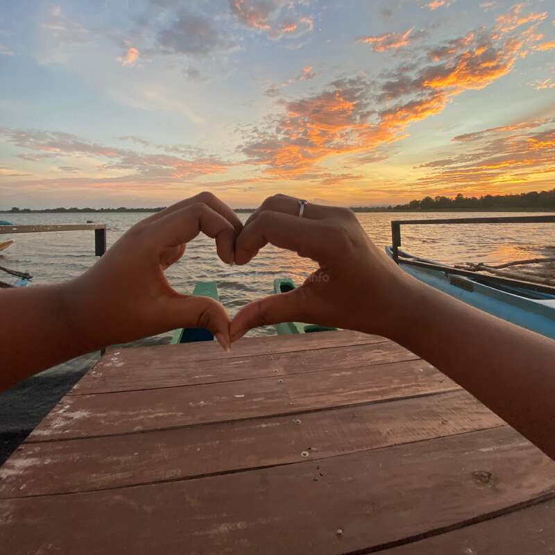 Dos manos forman un corazón sobre un hermoso cielo al atardecer, con vistas a un tranquilo lago con barcas, un muelle de madera y nubes dispersas iluminadas por una luz anaranjada.