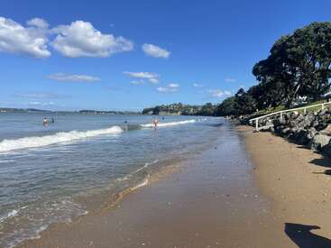 Une magnifique plage ensoleillée avec des vagues douces, des baigneurs, un ciel bleu limpide, des nuages cotonneux, un rivage sablonneux, des rochers et des arbres verdoyants le long du littoral.