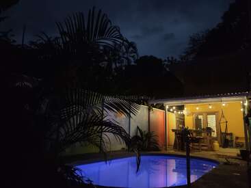 The image depicts a serene backyard scene at night, featuring a small pool, a covered patio with a table and chairs, and a yellow house with string lights.