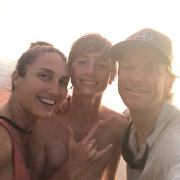 La imagen muestra a tres personas posando para un selfie en una playa, con el sol brillando a sus espaldas, creando un ambiente cálido y alegre.