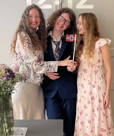 Tres personas con el pelo largo y rizado sonríen alegremente en el interior. Una de ellas sostiene una pequeña bandera noruega. Visten trajes de gala con motivos florales junto a un jarrón con flores.