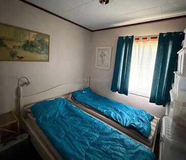 Una pequeña habitación con dos camas individuales cubiertas con mantas azules, una ventana con cortinas oscuras, dos cuadros en la pared y un mobiliario sencillo y minimalista. Ambiente acogedor.