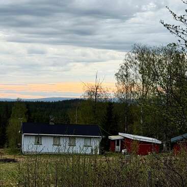 Uma cena rural com uma casa branca e um galpão vermelho, cercada por árvores e vegetação. O céu está nublado, com um toque de pôr do sol alaranjado no horizonte.