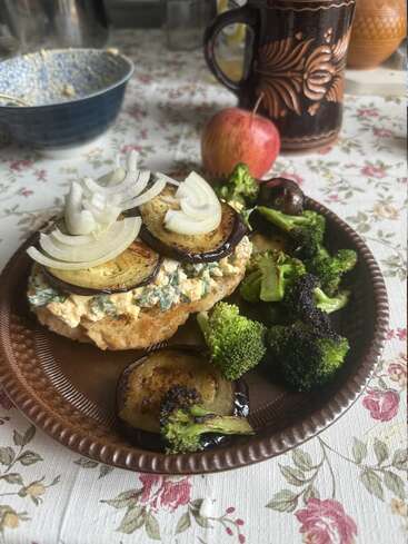Un sandwich rustique garni d'aubergines grillées, d'oignons et d'une pâte à tartiner crémeuse est servi avec des brocolis rôtis, une pomme, une tasse et une nappe fleurie.