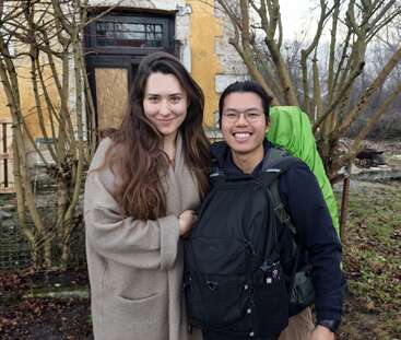 Une femme souriante vêtue d'un manteau beige se tient à côté d'un homme portant des lunettes et un sac à dos. Ils sont en plein air, à côté d'un bâtiment rustique et d'arbres dénudés.