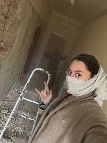 Une femme portant un foulard et se couvrant le visage prend un selfie pendant des travaux de rénovation. Des gravats, de la poussière, une échelle, des briques apparentes et une autre personne sont visibles derrière elle.