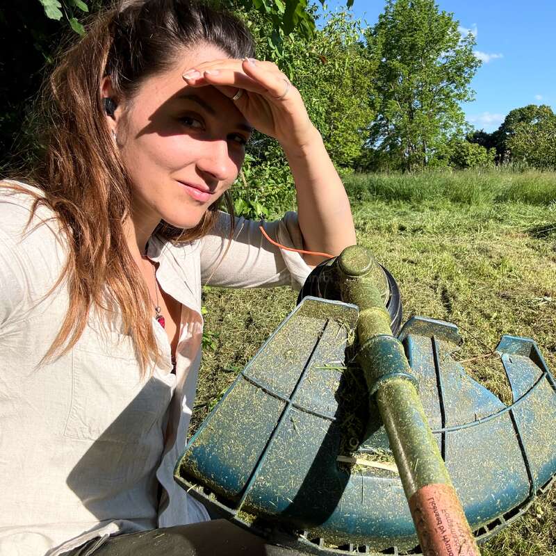 Une femme est assise à l'extérieur, en plein soleil, et se cache les yeux avec sa main. Elle tient un coupe-herbe, entourée d'herbe fraîchement coupée, et profite de la nature.