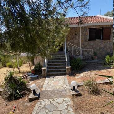 A stone house with a tiled roof is surrounded by dry landscaping, shrubs, and trees. A stone path leads to steps and a shaded entrance.