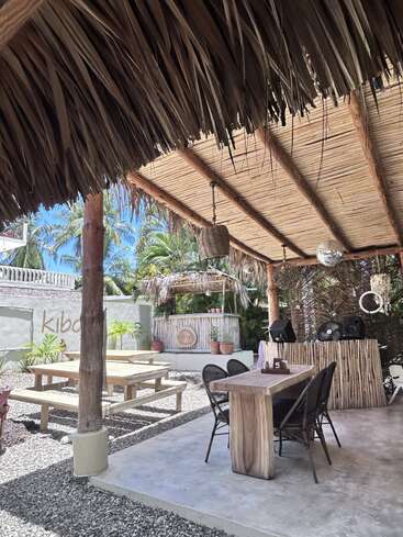 Dieses Bild zeigt ein tropisches Café oder eine Bar im Freien mit Strohdächern, Holztischen und -bänken, Topfpflanzen, Lautsprechern und zahlreichen Palmen unter strahlend blauem Himmel.