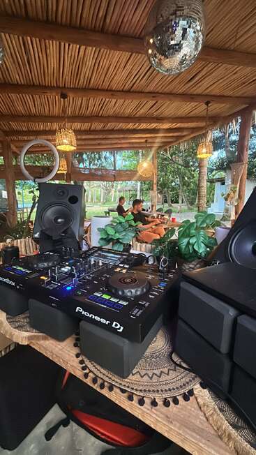 Eine gemütliche Außenanlage mit DJ-Pult, ausgestattet mit Pioneer-DJ-Equipment, Lautsprechern, viel Grün, entspannten Gästen, Holzdecke, Hängelampen und einer Discokugel über den Köpfen.