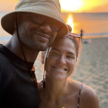 Ein lächelndes Paar posiert bei Sonnenuntergang am Strand für ein Selfie. Das warme Sonnenlicht spiegelt sich im Wasser und schafft eine friedliche, romantische und fröhliche Atmosphäre.